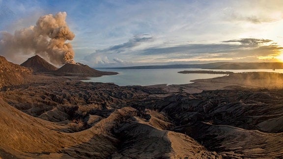 Panoramaaufnahme einer Insellandschaft, die von grauer Vulkanasche bedeckt ist. Ein Vulkan am linken Rand qualmt. 