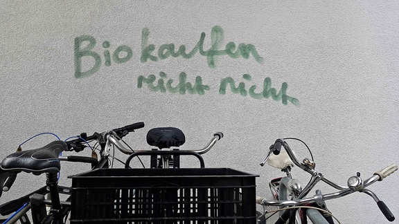 Grafitto: "Bio kaufen reicht nicht" steht an eine Wand geschrieben – davor teilweise sichtbar abgestellte Fahrräder