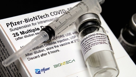 Eine Spritze und eine Impfstoffampulle liegen auf einer Schachtel mit dem Aufrduck: Pfizer-Biontech-Covid-19-Impfstoff.