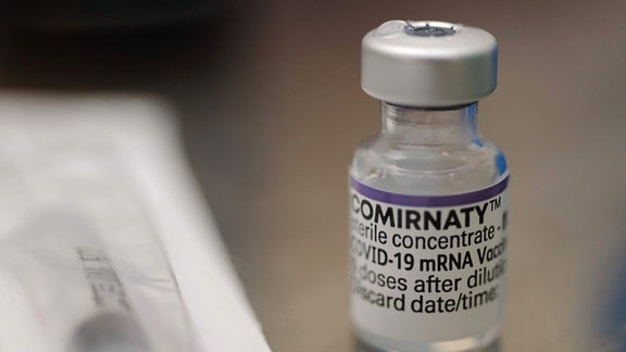 Ampulle mit dem Impfstoff Comirnaty von BioNTech. Zu sehen ist ein kleines Glasfläschchen mit dem grauen Deckel durch den die Impfkanüle eingestochen wird. Das Fläschchen hat ein weißes Ettiket mit einem lila Rand und der Aufschrift Comirnaty.