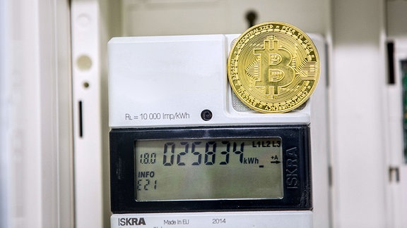 Eine goldfarbene Bitcoin-Münz-Attrappe steht auf einem Stromzähler, der die Zahl der Kilowattstunden im Display anzeigt.