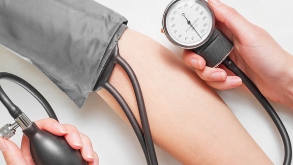 Ein Arm steckt in einem Blutdruckmessgerät, eine andere Hand hält die Luftpumpe und die Druckanzeige.