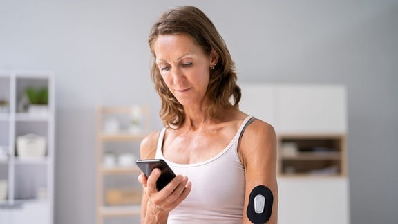 Eine Frau schaut auf ihr Handy, an ihrem Arm trägt sie einen Sensor für die Blutzuckermessung.