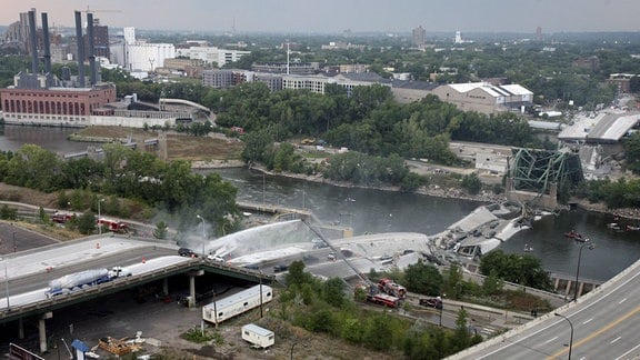 Luftbild Autobahnbrücke über Fluss in industriellem Vorortgebiet. Brücke und Fahrbahn auf Ufer und in Fluss gestürzt. Daneben Straße mit intakter Fahrbahn. 