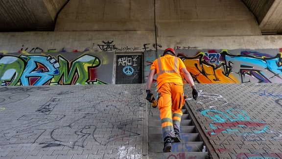 Person mit organgefarbenen Signalanzug läuft die Stufen am Pfeiler einer Autobahnbrücke hoch mit Gerät in der Hand, im Hintergrund Graffiti