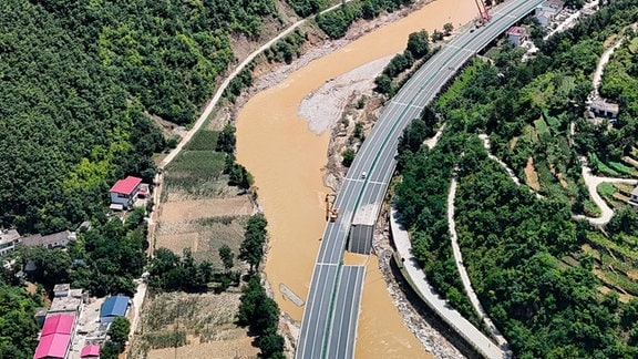 Luftbild: Teil der dreispurigen Seite einer sechsspurigen Straße in Flusstal ist in Fluss gestürzt, daneben Häuser und grüne Berge