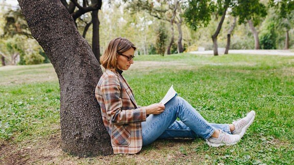 Eine Frau sitzt in einem Park an einen Baumstamm gelehnt und liest ein Buch.