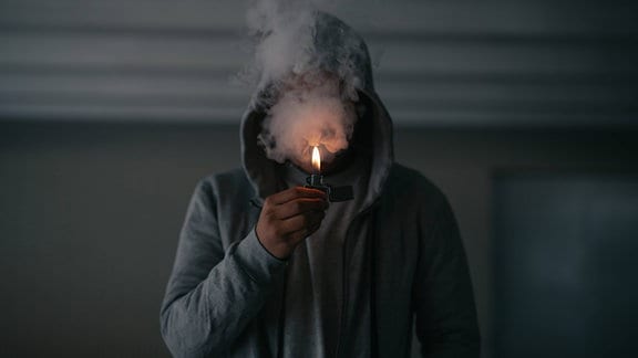 Nicht erkennbarer Mann in Kapuzenjacke, der Zigarette raucht, während er im dunklen Raum mit brennendem Feuerzeug in der Hand steht