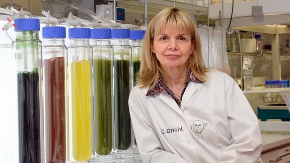 Eine Frau vor einer übergroßen Reihe an Reagenzröhrchen ´, die mit roten, grünen, gelebn Flüssigkeiten gefüllt sind