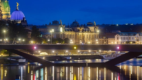 Ansicht von Fluss aus: Carolabrücke mit abgesacktem Teil im Wasser, im Hintergrund abendliche Altstadt mit farbig glänzenden Lichtern, die sich teilweise im Wasser spiegeln.