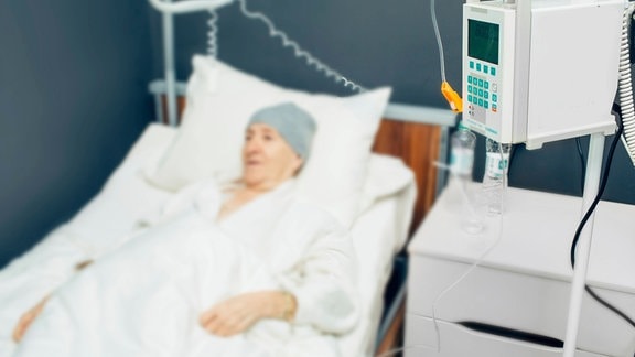 Eine ältere Person (unscharf im Hintergrund) erhält eine Chemotherapie. Sie liegt in einem weißen Krankenhausbett und trägt eine graue Wollmütze auf dem kahlen Kopf. Im Vordergrund stehen medizinische Geräte.