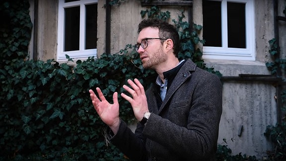Mann mit klein-kariertem Mantel, Drei-Tage-Bart und dickrandiger Brille in gestikulierender Erklärpose vor Hauswand mit Fenstern und Efeu