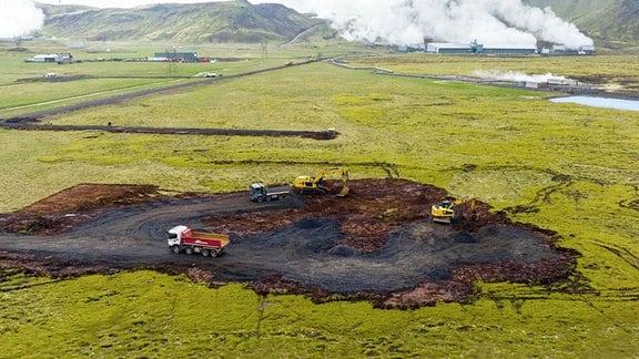 agger schaffen ein Baufeld auf Island. Im Hintergrund ist ein Geotheromie-Kraftwerk zu sehen, das von grauen Dampfwolken eingehüllt ist.