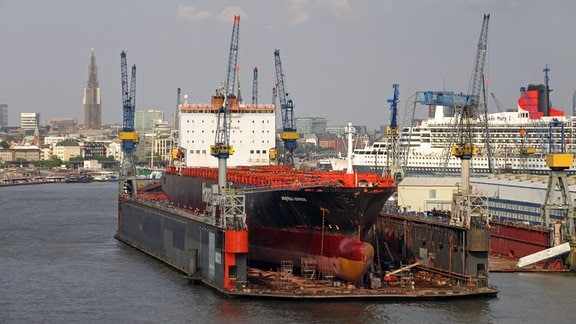 Das Containerschiff Montreal Express und das Kreuzfahrtschiff Queen Mary 2 in den Trockendocks von Blohm und Voss in Hamburg: Totale über eine hafenszenerie mit einem Dock über dem Wasser in dem ein großes Containerschiff steht.