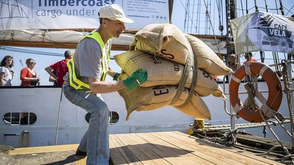Mann mit Basecap, Handschuhen und Weste befördert drei Säcke von einem Schiff, die an einem Seil hängen: Kaffee, Kakao, Rum und Kardamom werden von einem Segelschiff im Hamburger Hafen entladen.