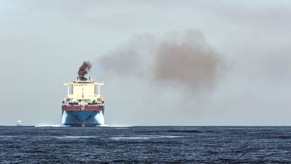 Ein Frachschiff auf dem Meer frontal am Horizont mit einer Rußwolke