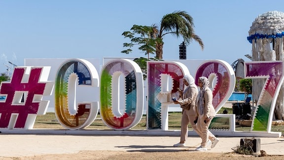 Auf sandigem Grund aufgestellte große bunte Buchstaben #COP27, sonniges Wetter, Palme im Hintergrund, Menschen laufen vor den höheren Buchstaben vorbei.