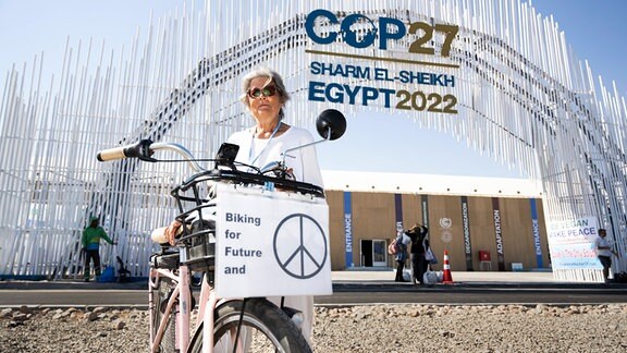 Ältere Frau mit kinnlangen grauen Haaren und Sonnenbrille seht mit E-Bike mit Aufschrift "Biking for Future and Peace" vor Torbogen mit Aufschrift "COP27".