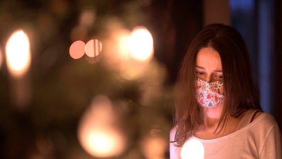 Eine Frau mit Mund-Nasenschutz-Maske schaut nachdenklich auf den leuchtenden Weihnachstbaum im Wohnzimmer