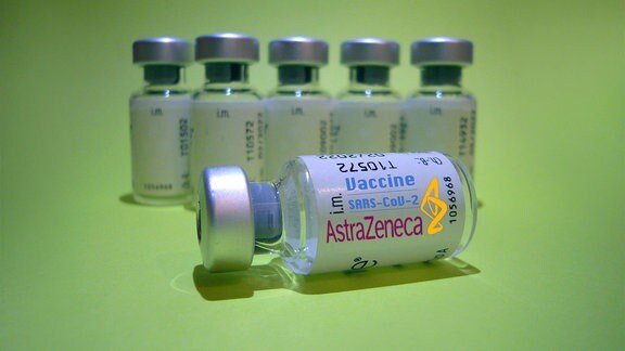 Symbolfoto: Kleine Glas-Behälter mit AstraZeneca-Etikett stehen aufgereiht vor grünem Hintergrund, eine liegt im Vordergrund.