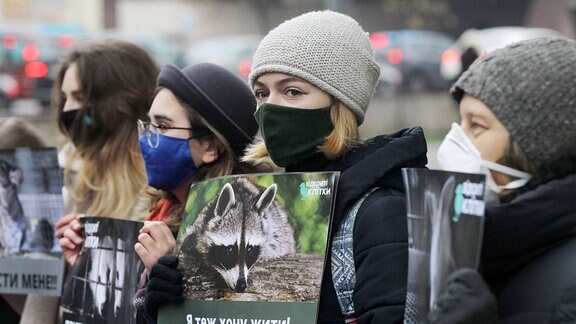 Junge Anti-Pelz-Aktivist/-innen mit Masken stehen perspektivisch schräg aufgereiht,  halten während der Demonstration Plakatee mit Text in kyrillischen Buchstaben und Fotos verschiedener Pelztiere hoch.