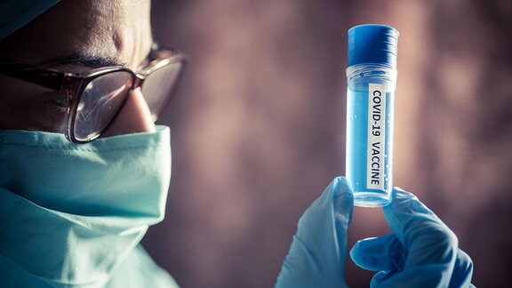 Person mit Brille (Gesicht im seitlichen Anschnitt) und Mund-Nasen-Schutz begutachtet einen Impfstoff mit Aufschrift "Covid-19 Vaccine", den sie in Gummihandschuhen hochhält. Cinematische, etwas mystische Farben, Hintergrund unscharf.