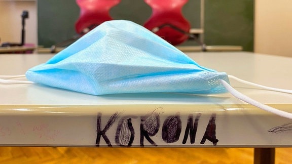 Mundschutzmaske und Schriftzug KORONA auf einem Tisch in einem leeren Klassenzimmer, im Hintergrund unscharf hochgestellte Stühle und Tafel