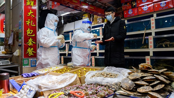 Zwei Menschen in weißen Schutzanzügen kontrollieren einen Lebensmittelstand und einen Händler auf einem Markt in China.