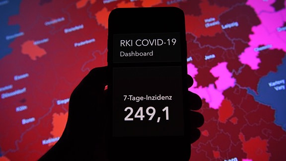 Eine Hand hält ein Handy hoch, auf dem Display steht: "RKI Covid-19 Deutschland: 7-Tage-Inzidenz 249,1". Im Hintergrund ist eine Deutschlandkarte mit Infektionszahlen zu sehen.