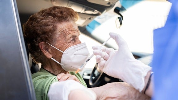 Ältere Frau mit FFP2-Maske auf Beifahrerinnensitz bekommt eine Impfung durchs Autofenster. Seitenansicht, impfende Person nur im Anschnitt-