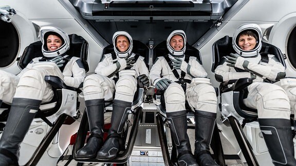Vier Raumfahrerinnen und -fahrer in auf vier Sitzen nebeneinander in weißen modernen Raumanzügen. Trainingssituation bei SpaceX.