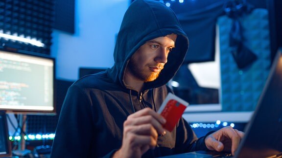 Ein Mann mit Kapuze sitzt vor einem Computer mit einer Chipkarte in der Hand. Düstere Stimmung, einfallendes Licht auf Gesicht.