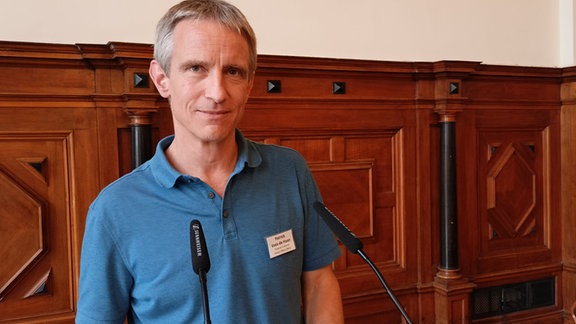 Patrick Voss-de Haan, ein Mann mit kurzen grauen Haaren und blauem Poloshirt steht an einem Rednerpult und schaut in die Kamera