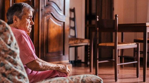 Seitenansicht in warmen Licht: Ältere Frau sind auf einem Sofa in einem Wohnzimmer und schaut leicht nach unten. Im Hintergrund Wohnzimmerschrank,, Tisch und Stühle aus Holz.