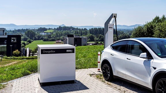 Eine weiße ChargeBox, ca. ein Kubikmeter groß, steht neben einem parkenden weißen Auto.