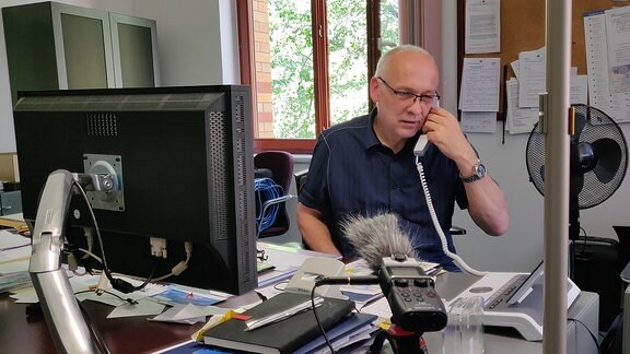 Mann mit Brille und kurzen, hellen Haaren in einem Büro am Telefon. Im Vordergrund Bildschirm und viele Dinge auf Schreibtisch.