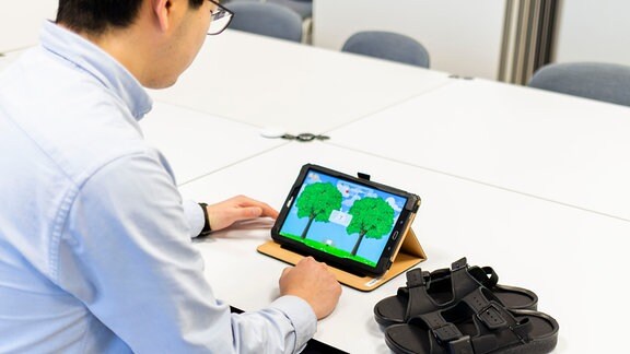 Mann sitzt mit Rücken zur Kamera an Tisch und trägt spezielle Birkenstock-ähnliche Sandalen. Auf Tisch Tablet-Computer mit Videospiel.