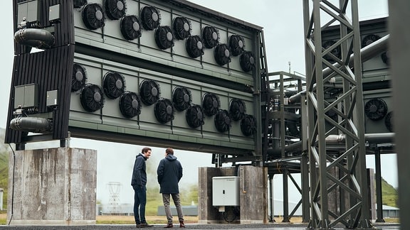 Industrieanlage: Direct Air Capture von der Rückseite. 24 Lüftungsventilatoren aufgereiht an einem Block, Rohre, darunter zwei Männer die sich mit Händen in den Taschen unterhalten.