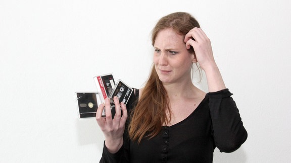Eine Frau schaut fragend auf zwei Kassetten und eine Diskette, die sie in ihrer rechten Hand hält.