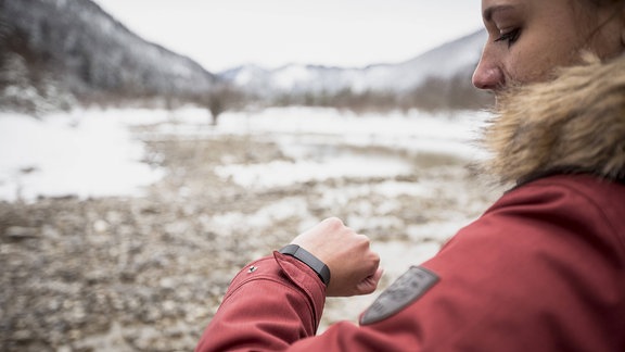 Frau in leicht verschneiter Gebirgslandschaft mit Bergen, Geröll und dicker roter Winterjacke mit Pelzkragen blickt auf Fitness-Tracker-Armband. Frau im Anschnitt rechts, Natur im Hintergrund unscharf.