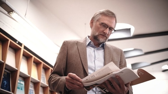Mann mit grauen Haaren, Brille, Bart und Anzug steht in einer Zeitschriften-Bibliothek, freundlich-interessierter Blick, blättert durch Zeitschrift, Blick von unten
