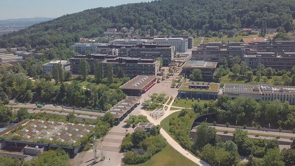 Drohnenaufnahme: Geländer der Universität Zürich (Irchel-Campus) von oben, mehrere wissenschaftliche Gebäude in einem grünen Park, dahinter grüner Hügel. 