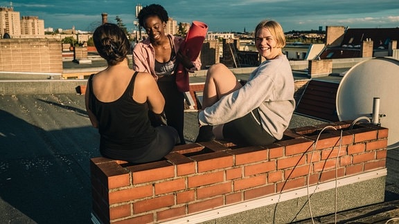 Drei fröhliche junge Frauen auf einem Dach in Berlin bei Sonnenauf- oder Untergang. Eine hält eine gerollte Yoga-Matte in der Hand, eine Sitze mit angewinkelten Beinen und schaut in Kamera, eine mit Rücken zur Kamera. Kleine Backstein-Mauer als Sitzgelegenheit, freundliches Wetter.