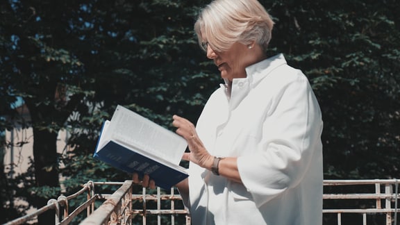 Frau mit hellen, kinnlangen Haaren und Brille und weißem Oberteil blättert auf einem Balkon in einem Buch, dunkelgrüner Baum bildfüllend unscharf im Hintergrund