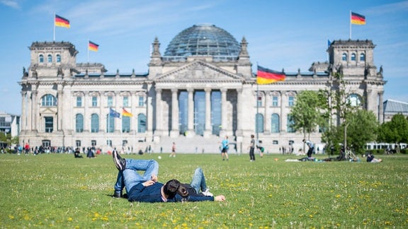 Reichstaggebäude in Berlin: Alter Bau mit hohen Säulen und Glaskuppel. Deutsche Flaggen auf Gebäude. Sonniges Wetter. Paar liegt auf Wiese beieinander und schaut auf Gebäude.