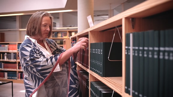 Frau mit kinnlangen, grauen Haaren und leichtem Poncho mit Muster. Seitenansicht, in Bibliothek, nimmt Buch aus Regal.