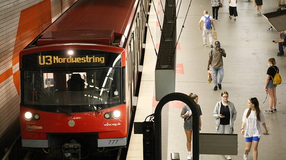 Draufsicht aus oberer Ebene: U-Bahn mit Zielanzeige "U3 Nordring" in U-Bahnhof. Abfahrtstafel an Decke, Menschen auf Plattform. Sitz in Fahrpersonal-Kabine, ohne dass eine Person dort sitzt.