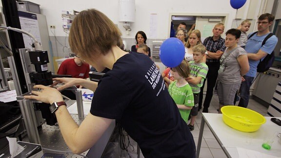 Eine Forscherin bedient ein Experiment, im Hintergrund stehen Zuschauer.