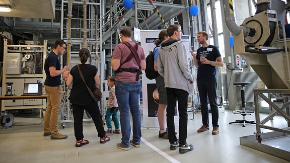 Besucher stehen in einer Technikhalle, in der dieverse Geräte in Stahlkonstruktionen eingebaut sind.