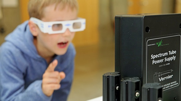 Ein Junge steht mit einer weißen Brille aus Pappe vor einem Gerät.